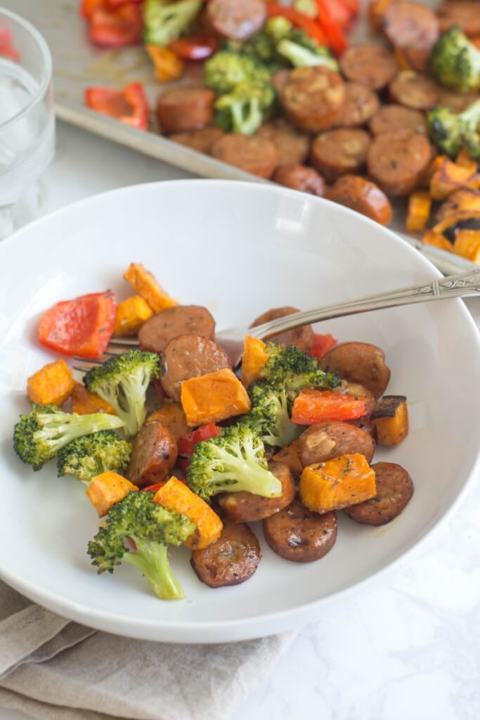 A white bowl of Sheet Pan Chicken Sausage & Veggies sits on a countertop with a brown linen napkin and a glass of water.